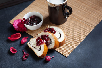 Slices of butter poppy roll, served with cherry jam. Large ceramic cup with hot drink. Black table and bamboo mat. Red dry flowers are scattered around. Ceramic mug has a beautiful decorative handle.