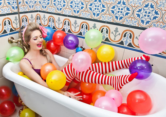Blonde woman with sunglasses playing in her bath tube with bright colored balloons. Sensual girl with white and red striped stockings having fun in bathroom, covered with balloons