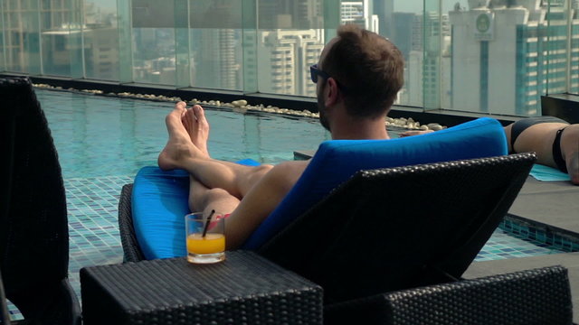 Young man lying on sunbed by pool on roof top in the city, super slow motion
