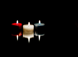 Red, white and blue lit candles on a mirror with reflections on a black background.