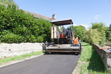 Fototapeta na wymiar Road tarring machine using a premix asphalt in a village tarring a narrow rural road viewed from behind showing the newly compacted tarmac