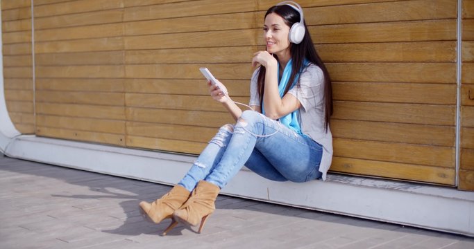 Attractive young woman listening to her music on stereo headphones as she sits on the ground in front of a closed kiosk