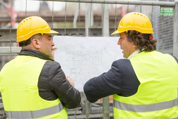 Back turned technician and architect at construction site reviewing office blueprints and technical drawings