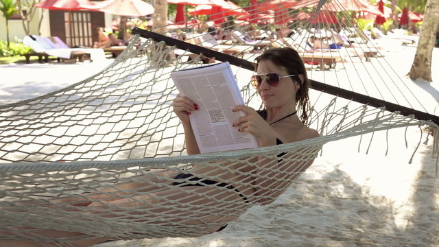 Young woman reading newspaper while lying on hammock, on the beach, 240fps

