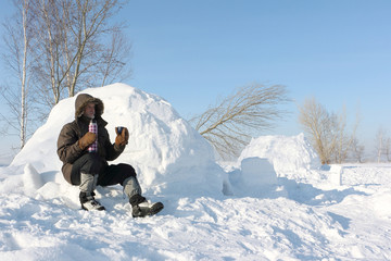 The man sitting near an igloo and drinking tea from thermos  in the winter
