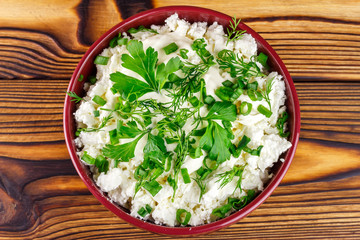 Fresh cottage cheese with sour cream, dill, parsley, onion in ceramic bowl on wooden table, top view