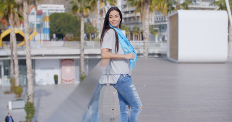 Fototapeta premium Single beautiful young smiling woman in long black hair ripped jeans and blue scarf looking back over glass railing overlooking park