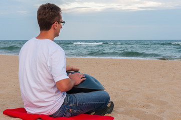 Young stylish guy sitting on the sand beach and playing handpan or hang with sea On Background. The...