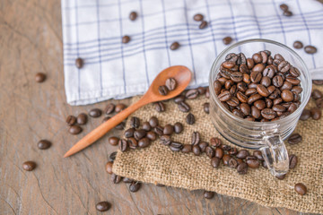 Coffee beans in a clear cup with wooden spoon are pile around of driftwood, sackcloth surface
