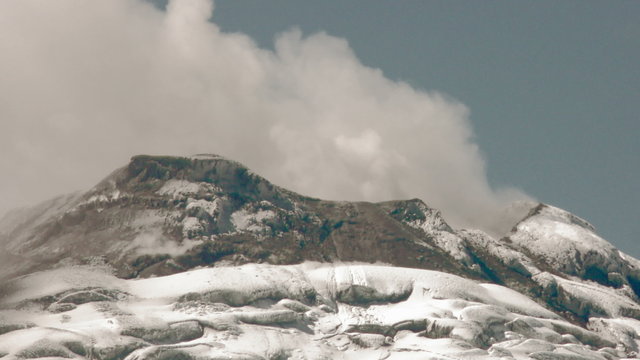 A majestic snow-covered peak of the Cotopaxi volcano with a glacier in the foreground in Ecuador, clouds drifting across the summit and slopes under a moody sky.