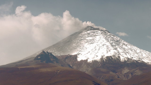 A majestic snow-covered peak of the Cotopaxi volcano rises above rugged terrain in Ecuador, clouds gently drifting around its summit in the high-altitude landscape.
