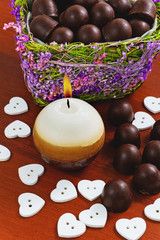 Chocolates in a box in the shape of a heart, a burning candle in the shape of a ball and white hearts on a wooden background