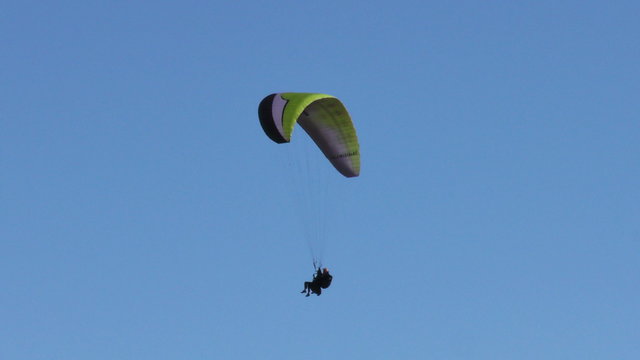 A paraglider with a green and black canopy glides through a clear blue sky above white clouds with a mountain peak behind near the Tungurahua volcano in Ecuador, a low-angle view.