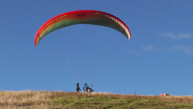 A paraglider with a red and green canopy descends and lands on a grassy hilltop, assisted by people on the ground near Banos, Ecuador, the approach, touchdown and canopy deflation.