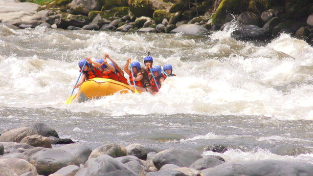 A group in a yellow raft navigate turbulent whitewater rapids on a rocky river near Banos, Ecuador, paddling together with a guide at the back in a forested river environment with large boulders.