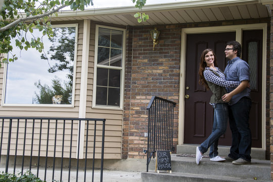 New Home Owners On Front Door Steps Of New Home As Dog Looks Out Window