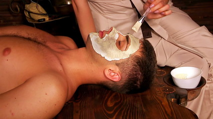 Man lying on a wooden massage table with clay facial mask in beauty spa. Used tripot.