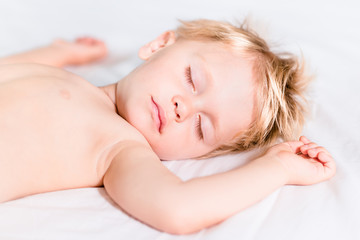 Close-up portrait of cute little kid with blond hair sleeping on white bed. Carefree childhood concept