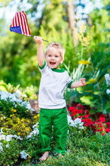 Barefoot little boy with blond hair laughing and waving american flag in sunny park or garden on summer day. Portrait of child on blurred background. Independence Day, Flag Day concept