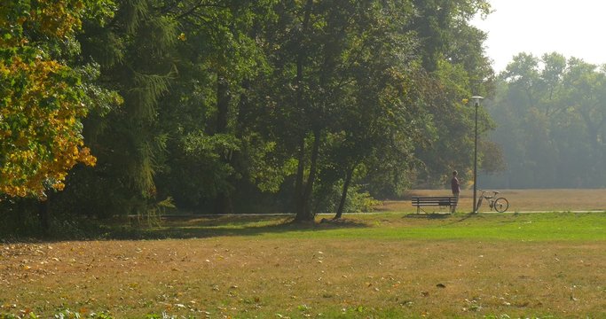 Man is Standing at The Bench Walking Man Has Stratched Took His Backpack from the Bench Bicycle is Standing at the Street Lamp in Park Wide Shot Sky