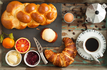 Breakfast flat lay of freshly baked bread, croissant, pretzel roll along with fruit, boiled egg and coffee. All served on a rustic wooden tray table.