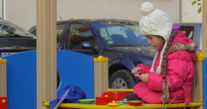 Little Blonde Girl With Braid is Playing Cook Toy Plates on a Table with Sand Toy Dishes Toy Food Forks Spoon Girl is Playing in the Sandbox Outdoors