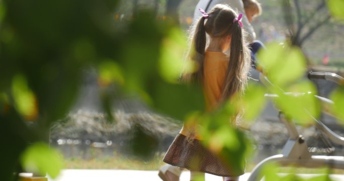 Little Girl With Long Fair Hairs in Orange T-Shirt is Running by Playground to the Swing Children Playing at Playground View through the Green Leaves