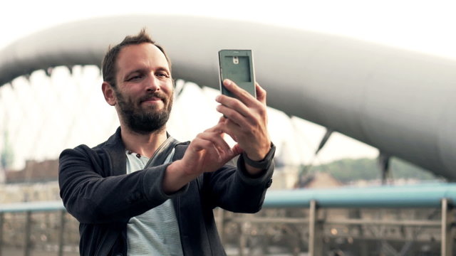 Man taking selfie photo with cellphone, standing by the bridge
