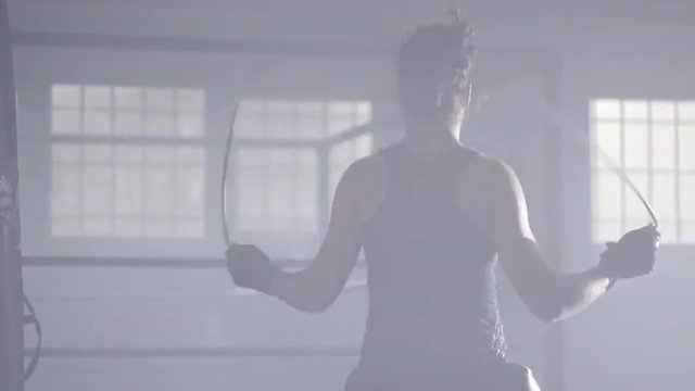 Female athlete skipping with her back towards camera, in a boxing gym with a ring at background. Dolly move. UHD, 4K.