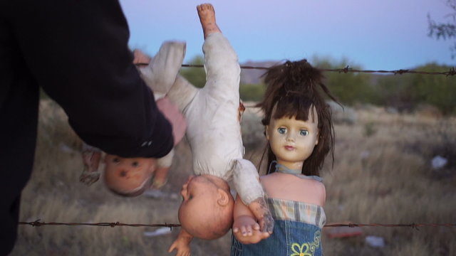 Static shot of a mentally unstable or crazy person hanging toy dolls on a barbed wire fence and grabbing the neck in a motion of strangling it.