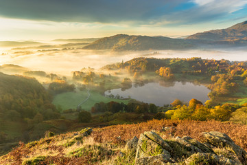 Fototapeta premium Beautiful Autumnal fog lingering over Windermere valley with Loughrigg Tarn in foreground. 
