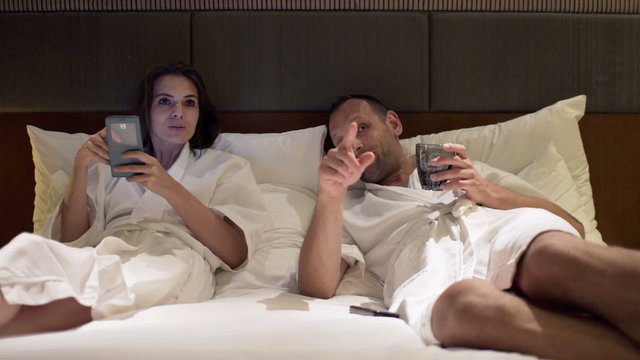 Young couple watching tv and using smartphone in bed during night

