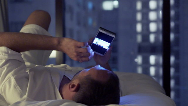 Attractive, young man browsing photos on smartphone lying in bed at night 
