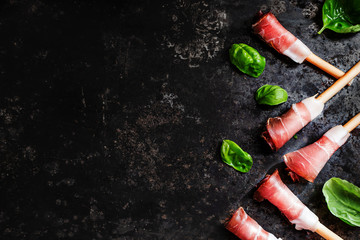 Grissini bread sticks with prosciutto ham and basil over black metal surface