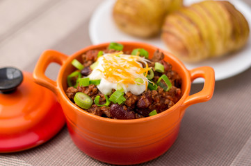 Fresh homemade bowl of chili con carne, with beans, sour cream, grated cheddar cheese and green onions, served with hasselback potatoes on the side.