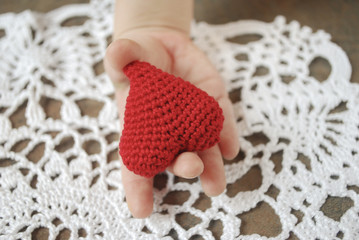 Child's hand holding crochet red heart on white napkin background.