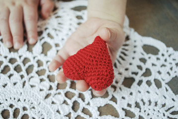 Child's hand holding crochet red heart on white napkin background.