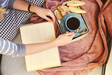 Female hands with open book, plaid, fruits and coffee on sofa. Top view