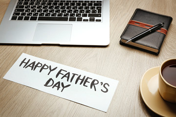 Happy Father's Day inscription with laptop, notebook and coffee cup on wooden background. Greetings and presents