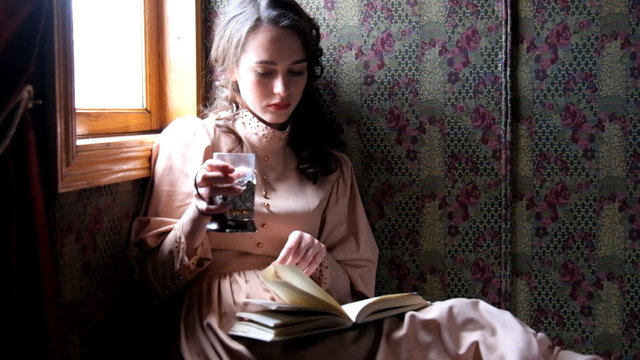 Young woman dressed in retro dress sitting in old train compartment and reading book drinking tea.