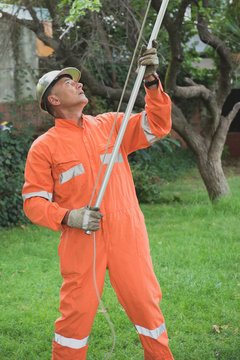 Adult worker wearing an orange safety suit and helmet. He is pruning and measuring trees.