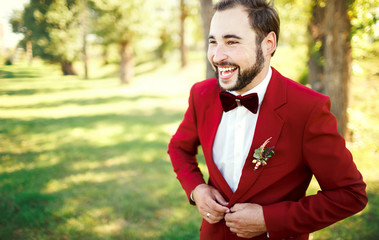 Stylish groom in tuxedo laughs suit marsala red, burgundy bow tie. Man buttoning his jacket outdoors. Professional hairstyle, beard, mustache. Wedding preparations, getting ready. Copy space for text.