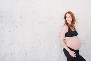 Pregnant woman with red hair in black clothes on a white wall