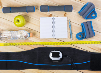 Electrical Muscle Stimulation Belt, Dumbbells, Ankle Weights, Tape Measure, Apple, Bottle Of Water, Notepad To Workout Or Diet Plan On Wooden Floor. Sport Fitness Background.