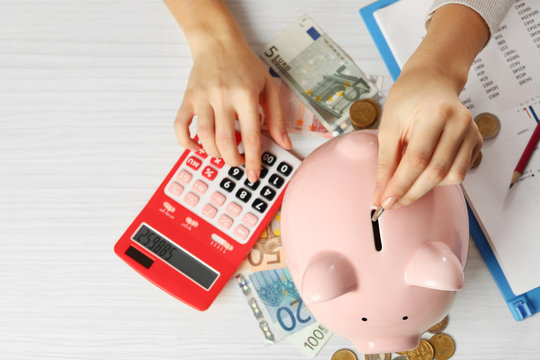 Woman's hands putting euro coin into a piggy bank and counting with calculator on the table. Financial savings concept