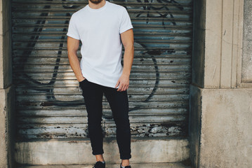 Bearded guy wearing white blank t-shirt and black jeans, standing opposite garage 