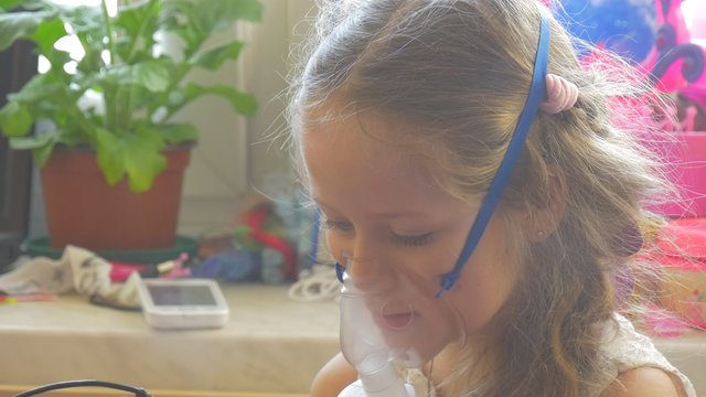 Girl Kid is Breathing Through Inhaler Playing with Mobile Phone Sitting With Nebulizer Mask on Her Face Holding a Mobile Phone Steam is Raising Up