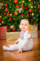 Cute little baby girl sitting in front of Christmas tree with colorful lights and a lot of gift boxes