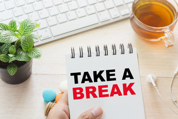 Man holding TAKE A BREAK message on book and keyboard with a hot cup of tea, macaroon on the table. Can be attributed to your ad.