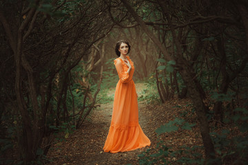 Fototapeta premium Piękna hrabina w długiej pomarańczowej sukience spaceruje po zielonym lesie pełnym gałęzi, elfka, księżniczka w sukience vintage, królowa lasu, modne stonowane kreatywne kolory komputerowe
